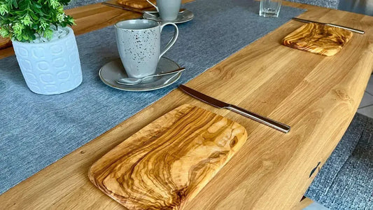 Large set wooden table with coffee cups, knives, a small potted plant, and 22 cm olive wood breakfast boards at each place setting